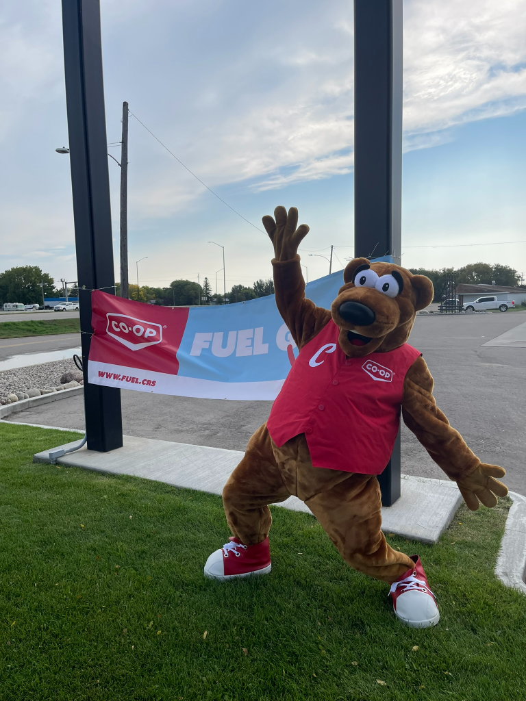 Co-op Cooper bear mascot posing in front of a Fuel Good Day banner outdoors near a Co-op fuel site.