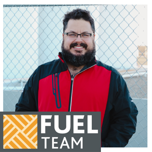A smiling Valleyview Co-op team member stands outdoors on a sunny day. He is wearing a red and black jacket, with his hands in the jacket pockets. The bottom of the image shows a Fuel Team logo, with a yellow and grey geometric design.
