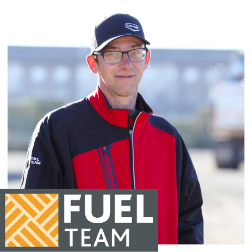 A smiling Valleyview Co-op team member stands outdoors on a sunny day. He is wearing a red and black jacket, and a black hat featuring the Co-op logo. The bottom of the image shows a Fuel Team logo, with a yellow and grey geometric design.